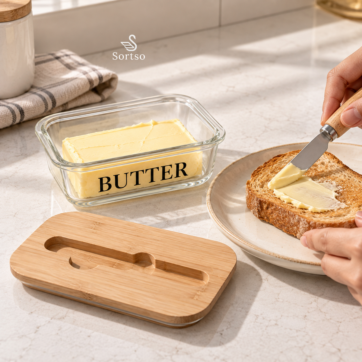 Glass butter keeper with bamboo lid and knife being used to spread butter on toast